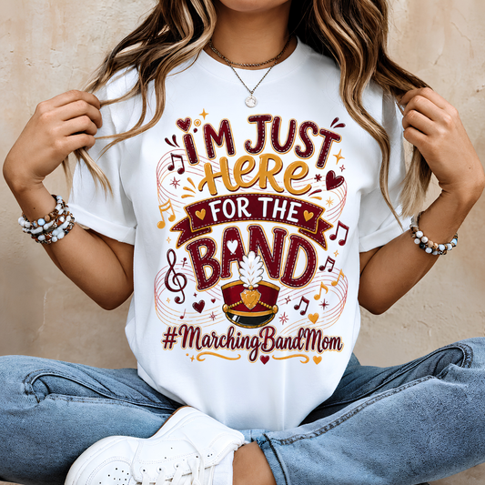 A woman wearing a white graphic t-shirt with a 'Marching Band Mom' design, paired with blue jeans and white sneakers, sitting against a neutral beige wall.